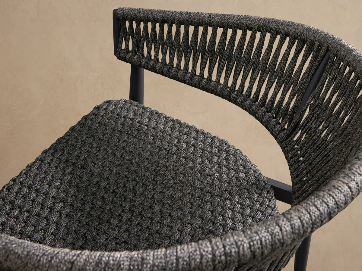 Front view of Talia Outdoor Aluminum Lounge Chair, dark gray woven rope back and seat, charcoal aluminum frame, on textured floor.