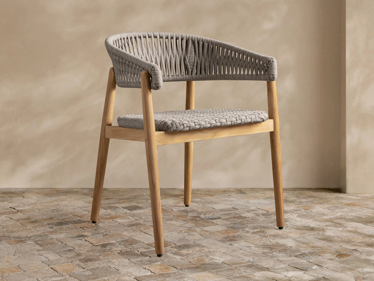 Talia Outdoor Teak Dining Chair, golden teak frame with grey woven rope seat and rounded back, angled on rustic tiled floor.