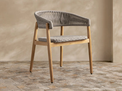 Talia Outdoor Teak Dining Chair, golden teak frame with grey woven rope seat and rounded back, angled on rustic tiled floor.