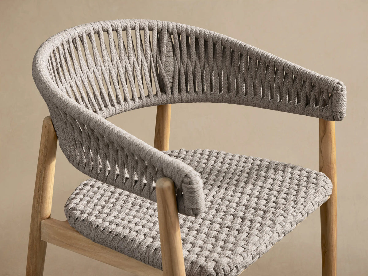 Talia Outdoor Teak Dining Chair, front view. Natural golden teak frame supports a curved light gray woven rope backrest & woven seat. Tiled floor.