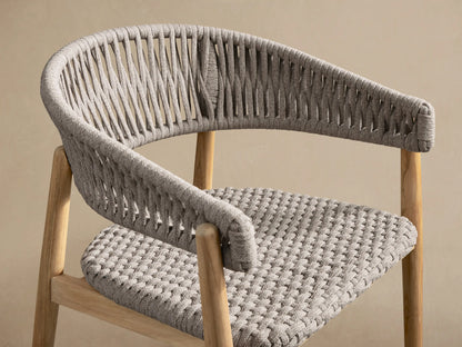 Talia Outdoor Teak Dining Chair, front view. Natural golden teak frame supports a curved light gray woven rope backrest & woven seat. Tiled floor.