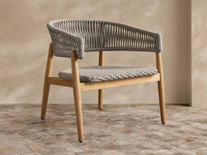 Talia Outdoor Teak Lounge Chair. Light brown teak wood frame with gray woven rope back, arms, and braided seat, on stone floor.