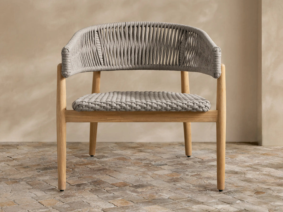 Talia Outdoor Teak Lounge Chair. Light brown teak wood frame with gray woven rope back, arms, and braided seat, on stone floor.