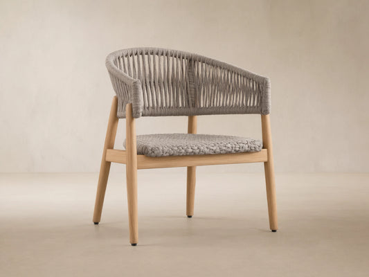 Talia Outdoor Teak Lounge Chair. Light brown teak wood frame with gray woven rope back, arms, and braided seat, on stone floor.