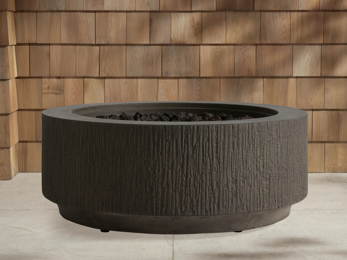 Wilmar Round Natural Gas Fire Pit, dark gray concrete with vertical ribbed texture, lit flames over lava rock, on patio with wooden wall.