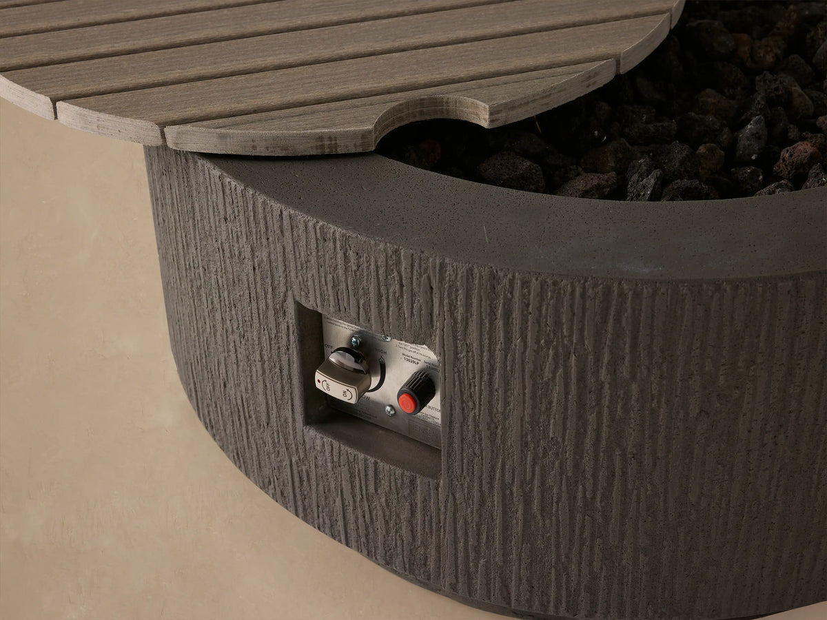 Wilmar Round Natural Gas Fire Pit. Dark gray concrete, round, with vertical textured sides & a slatted lid, on a tan background.