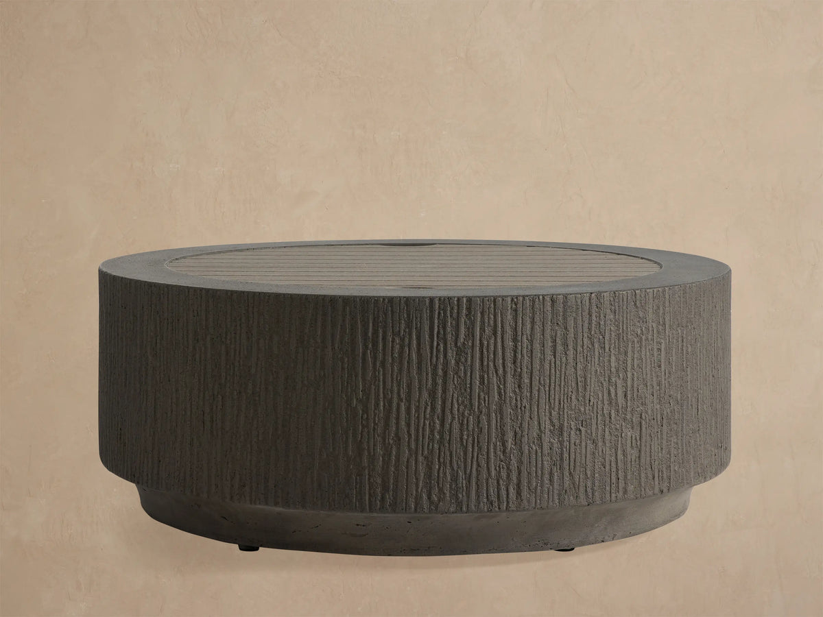 Dark gray Wilmar Round Propane Fire Pit, textured concrete with vertical grooves, filled with black lava rock, on a light patio.