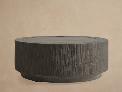 Dark gray Wilmar Round Propane Fire Pit, textured concrete with vertical grooves, filled with black lava rock, on a light patio.