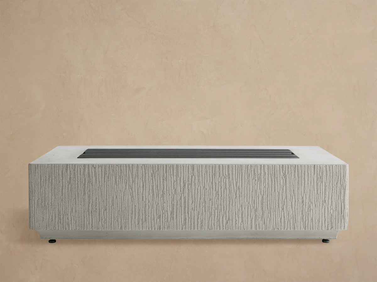 Rectangular Wilmar Fire Pit in light gray concrete, vertically textured, filled with black lava rocks, against a wooden shingle wall.