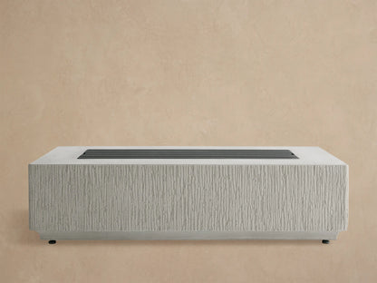 Rectangular Wilmar Fire Pit in light gray concrete, vertically textured, filled with black lava rocks, against a wooden shingle wall.