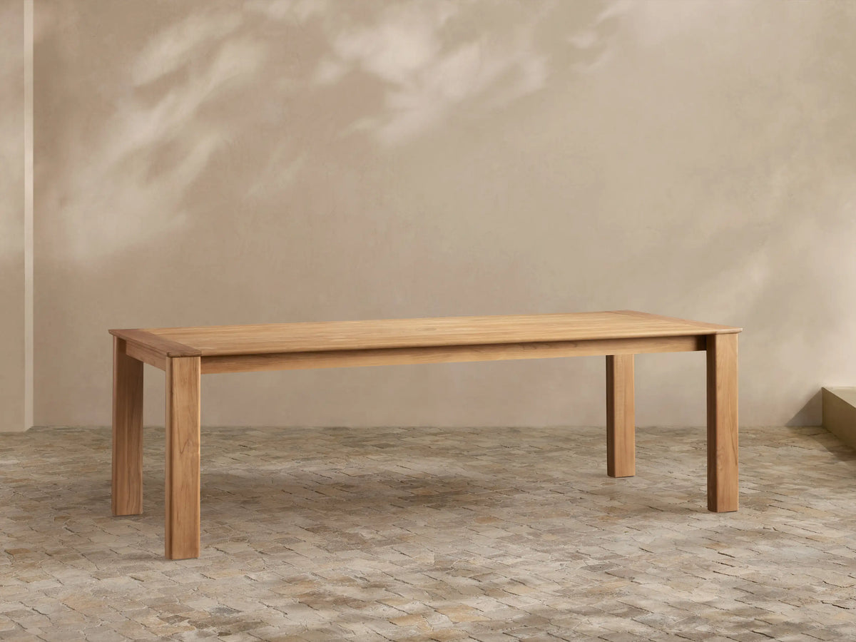 Zoe Outdoor Dining Table, rectangular natural teak wood, sturdy legs, on a stone patio with plant shadows on a light wall.
