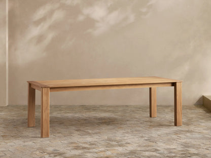 Zoe Outdoor Dining Table, rectangular natural teak wood, sturdy legs, on a stone patio with plant shadows on a light wall.