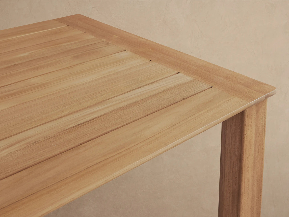 Rectangular Zoe Outdoor Dining Table made of natural teak wood, against a light wall with shadow patterns, on a paved floor.