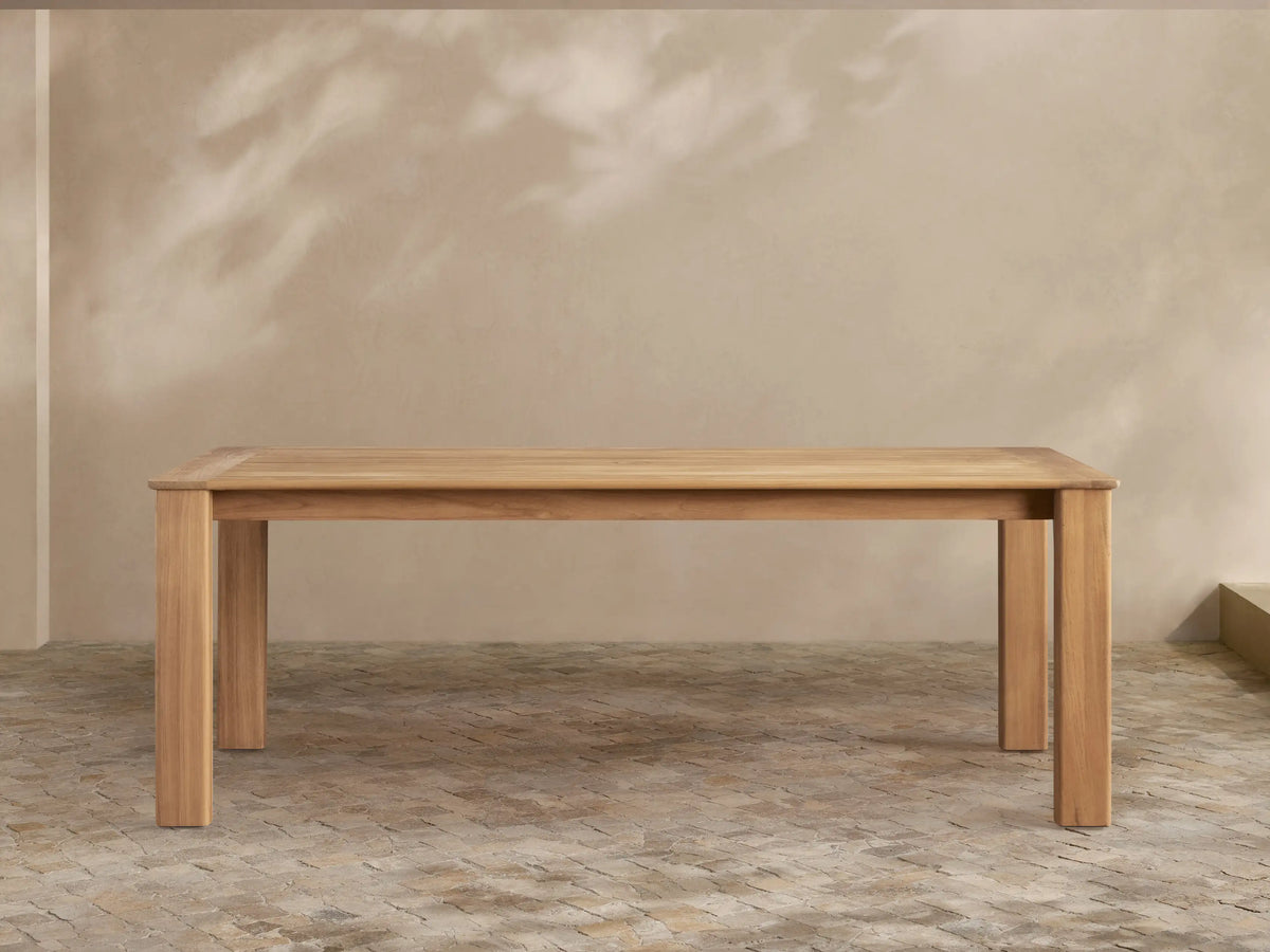 Zoe Outdoor Dining Table: Rectangular, golden-toned natural teak wood with sturdy legs, on a patterned patio floor.