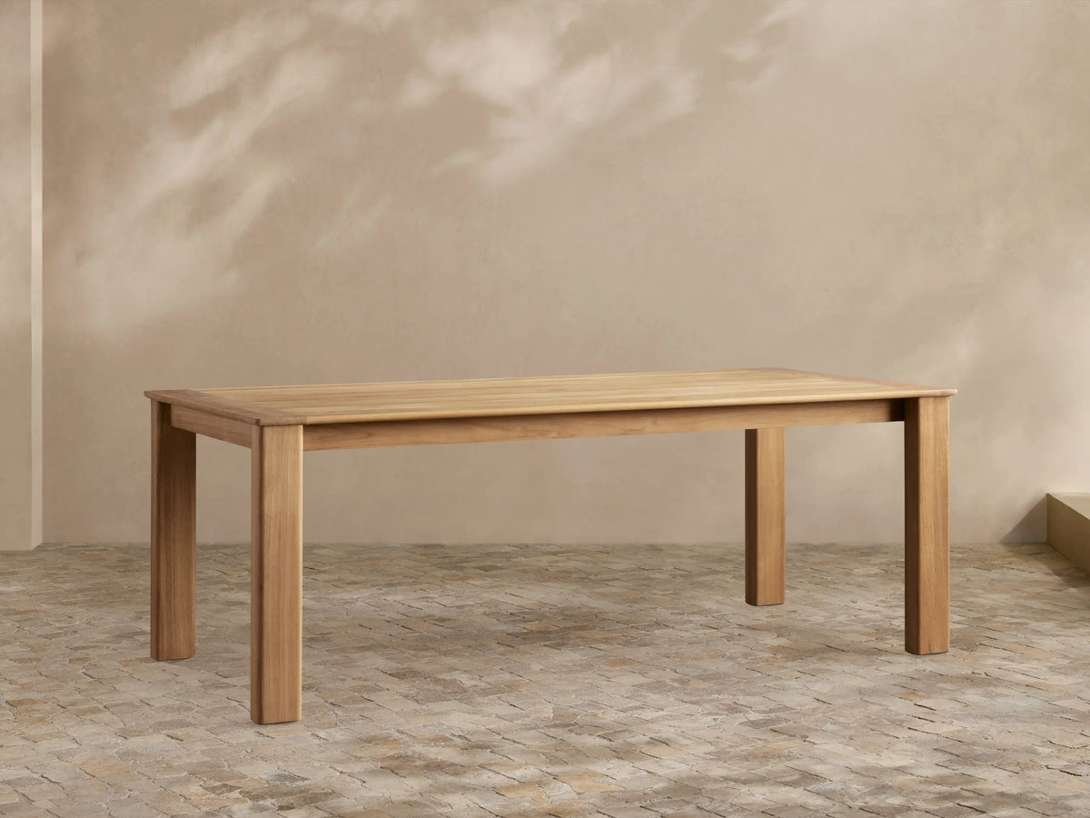 Zoe Outdoor Dining Table: Rectangular, golden-toned natural teak wood with sturdy legs, on a patterned patio floor.