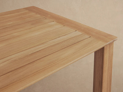 Zoe Outdoor Dining Table, rectangular teak wood with a slatted top and sturdy legs, on a stone patio against a light wall.