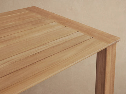 Rectangular Zoe Outdoor Dining Table made of natural golden-brown teak wood, on a tiled patio against a light wall.