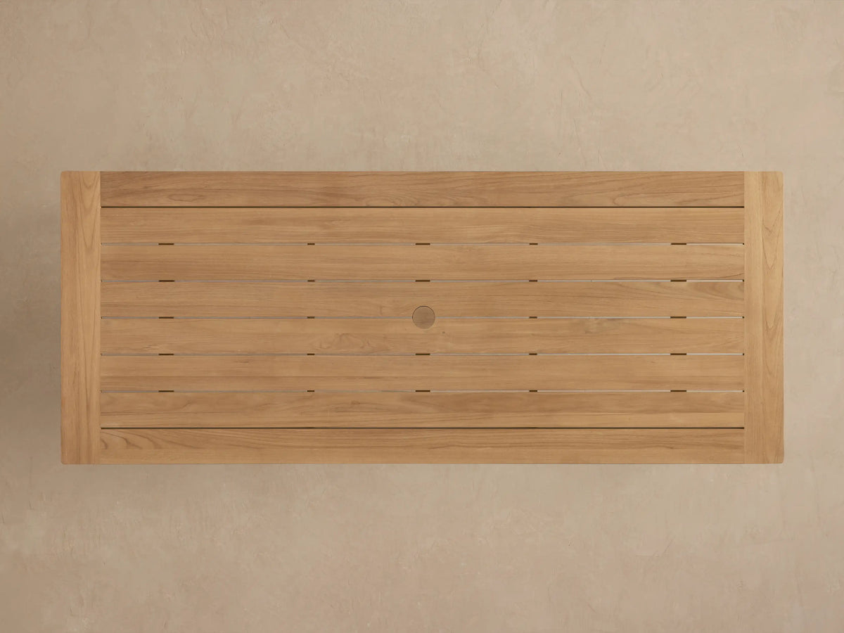 Angled close-up of Zoe Outdoor Dining Table. Golden-brown teak wood slat top with visible grain, sturdy leg.