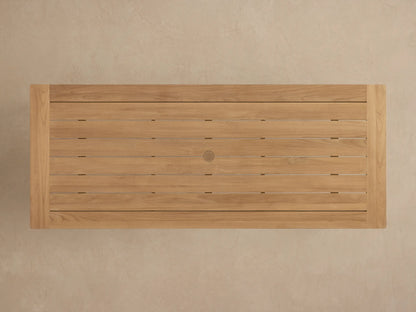Angled close-up of Zoe Outdoor Dining Table. Golden-brown teak wood slat top with visible grain, sturdy leg.