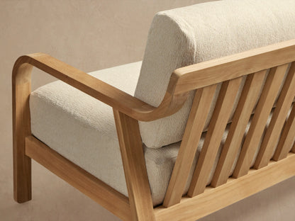Zoe Outdoor Sofa, light natural teak frame with curved arms, three off-white seat & back cushions, on a stone patio.