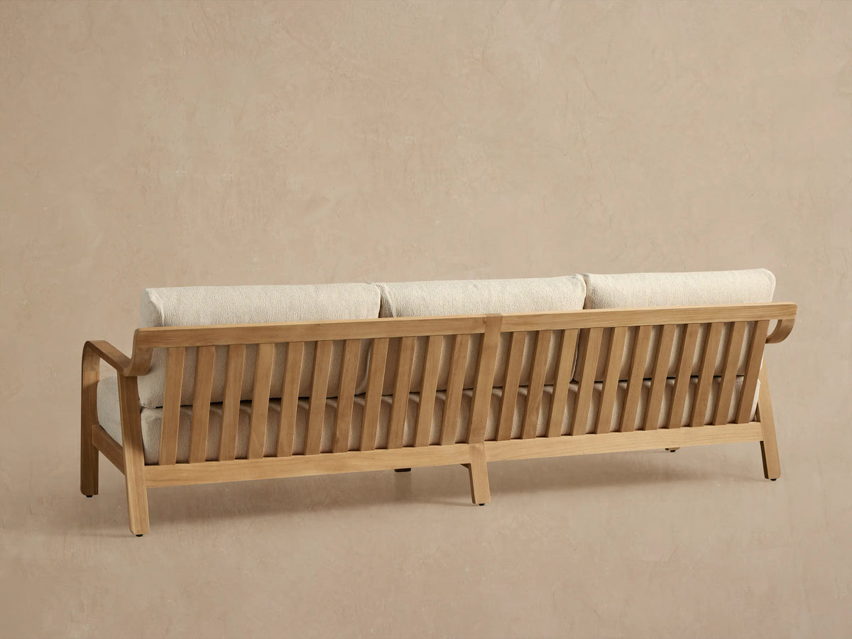 Angled view of a Zoe Outdoor Sofa with natural teak wood frame, slatted back, and light beige textured cushions.