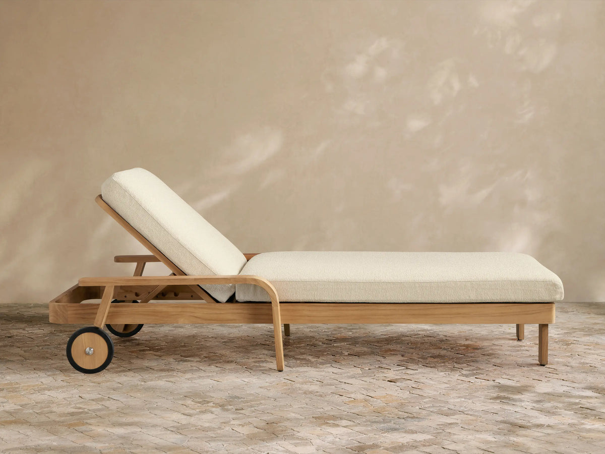 Zoe Outdoor Chaise, natural teak wood frame with cream cushions, two wheels on left, on a stone patio, angled against a light brown wall.