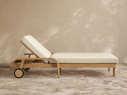Zoe Outdoor Chaise, natural teak wood frame with cream cushions, two wheels on left, on a stone patio, angled against a light brown wall.