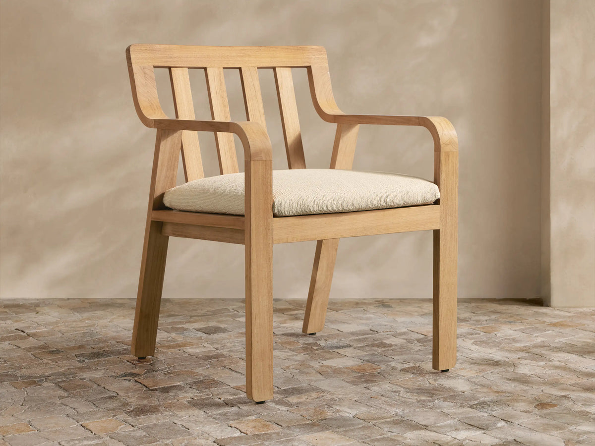 Zoe Outdoor Dining Arm Chair, light natural teak with slatted back, curved arms, and cream seat cushion on a stone floor.