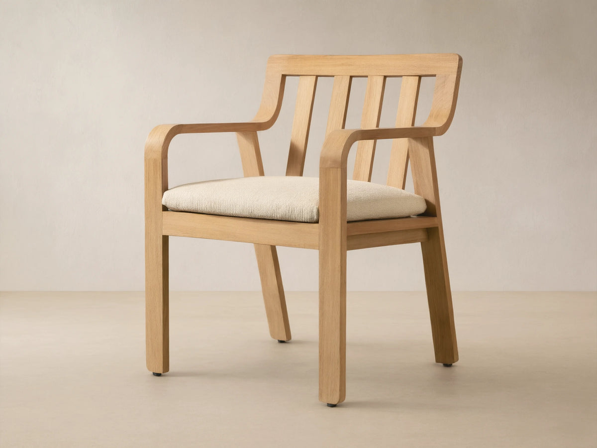 Zoe Outdoor Dining Arm Chair, light natural teak with slatted back, curved arms, and cream seat cushion on a stone floor.