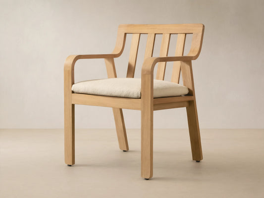 Zoe Outdoor Dining Arm Chair, light natural teak with slatted back, curved arms, and cream seat cushion on a stone floor.