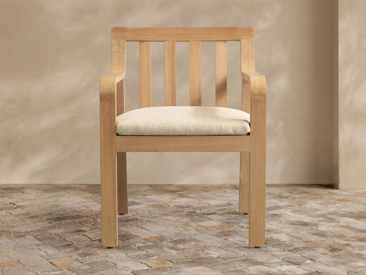 Zoe Outdoor Dining Arm Chair, light natural teak with slatted back, curved arms, and cream seat cushion on a stone floor.