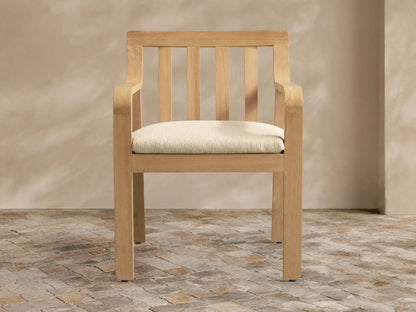Zoe Outdoor Dining Arm Chair, light natural teak with slatted back, curved arms, and cream seat cushion on a stone floor.