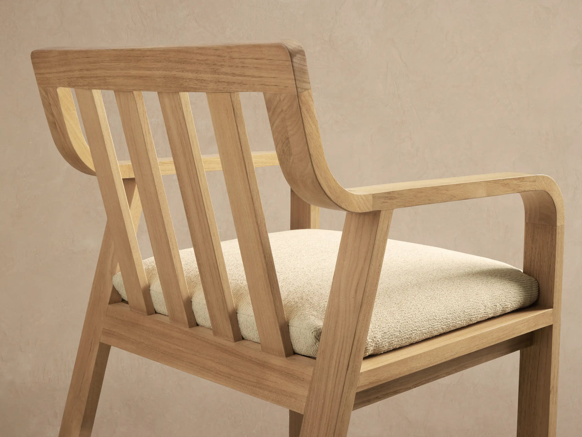 Front view of a Zoe Outdoor Dining Arm Chair in light natural teak wood with a slatted back and cream seat cushion, on a stone floor.