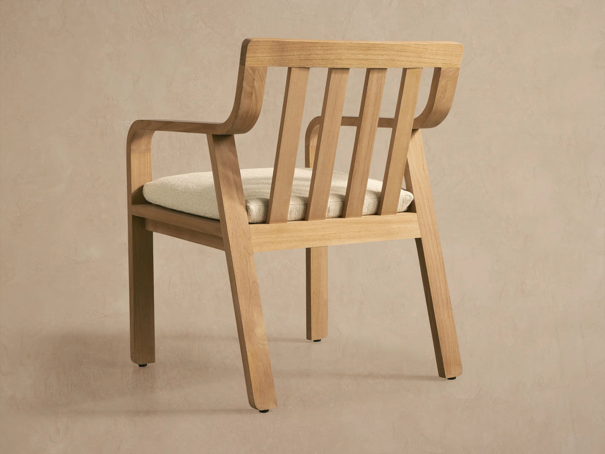 Zoe Outdoor Dining Arm Chair, natural teak wood, slatted back, curved arms, light beige textured seat cushion.