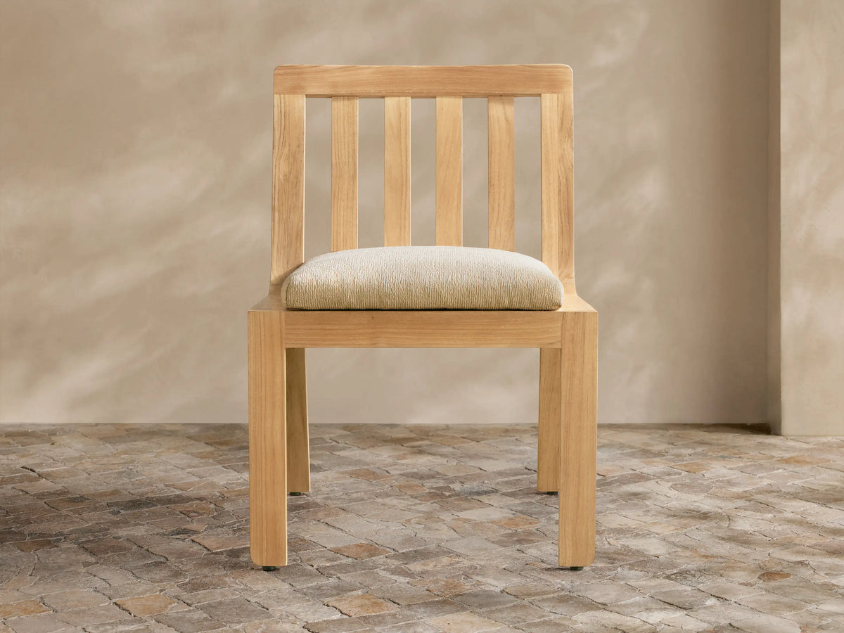 Natural golden teak Zoe Outdoor Dining Side Chair with slatted back, light beige cushion, on tiled floor.