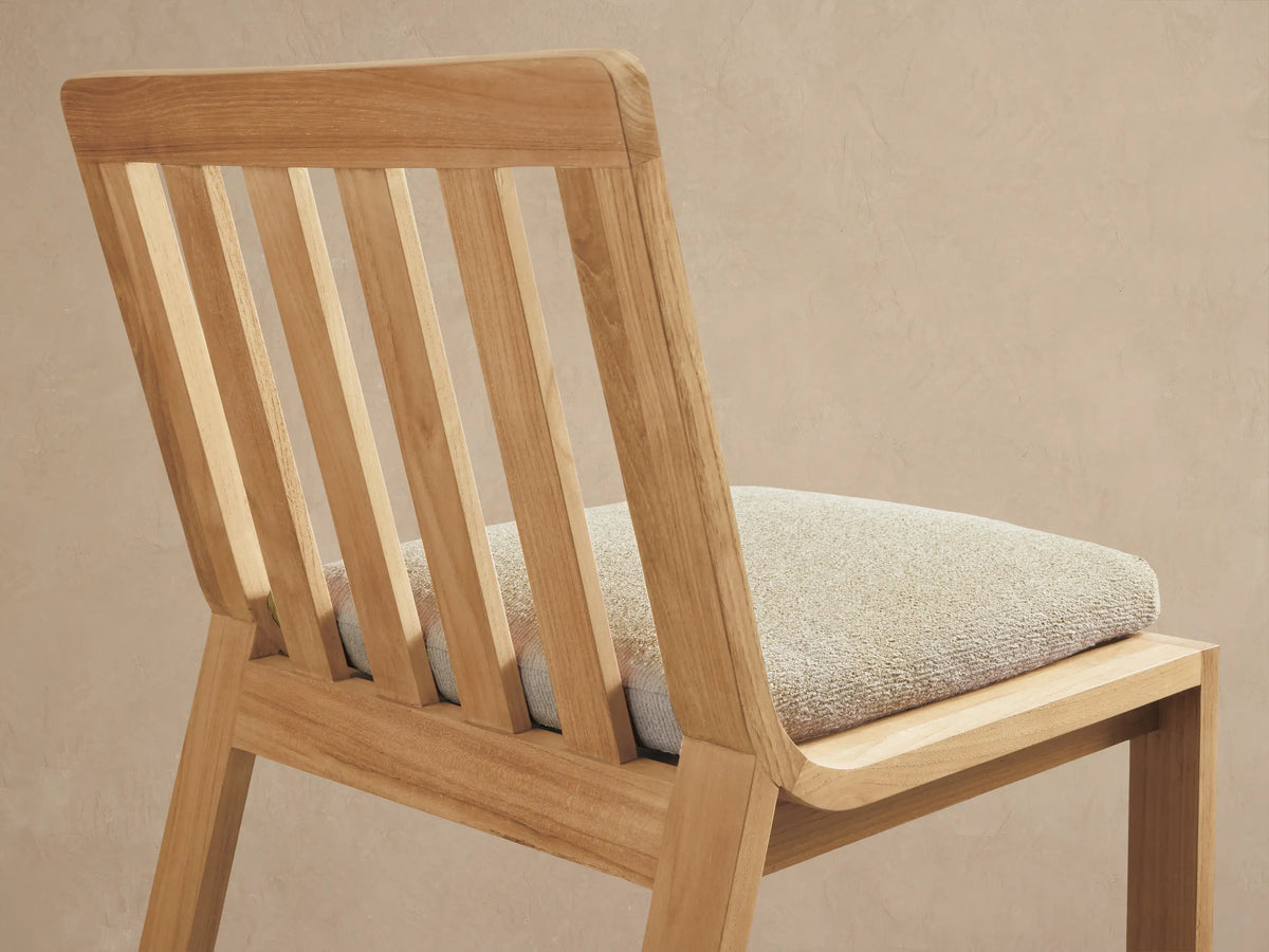 Zoe Outdoor Dining Side Chair, natural golden-brown teak wood with slatted back and light beige seat cushion, angled back view.