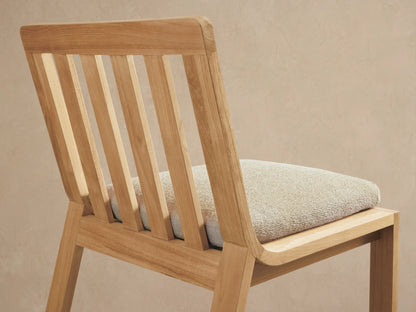 Zoe Outdoor Dining Side Chair, natural golden-brown teak wood with slatted back and light beige seat cushion, angled back view.