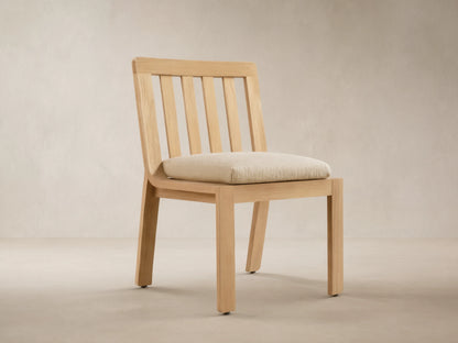 Natural golden teak Zoe Outdoor Dining Side Chair with slatted back, light beige cushion, on tiled floor.