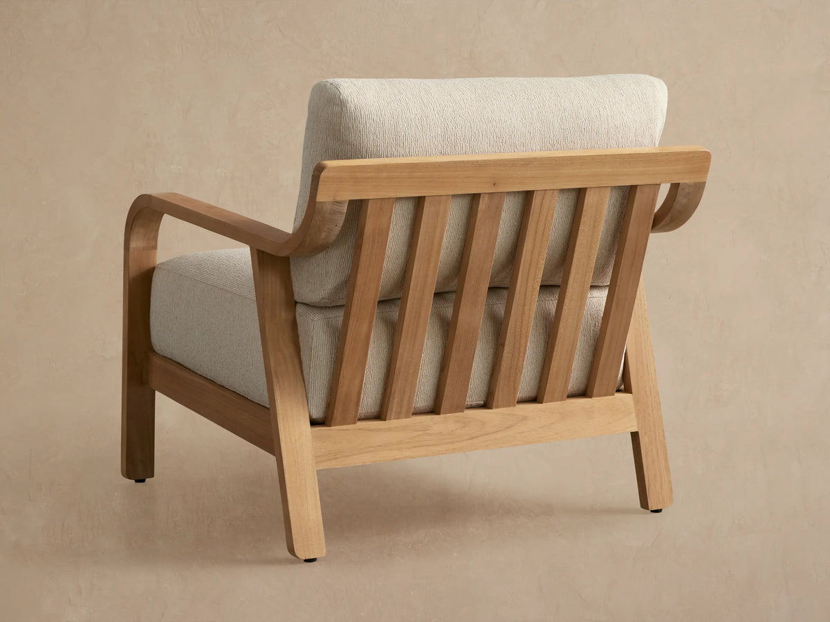 Angled close-up of Zoe Outdoor Lounge Chair. Golden teak wood frame with curved armrest, slatted back, and cream textured cushions.