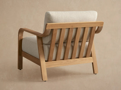Angled close-up of Zoe Outdoor Lounge Chair. Golden teak wood frame with curved armrest, slatted back, and cream textured cushions.