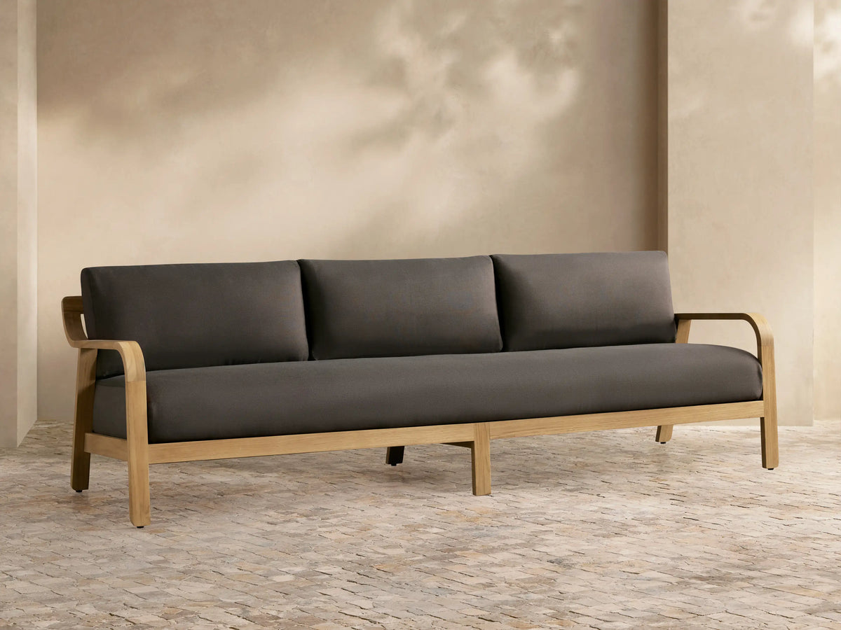 Front view of the Zoe Outdoor Sofa with dark grey cushions and teak frame on a patio, next to a striped armchair and stone coffee table.