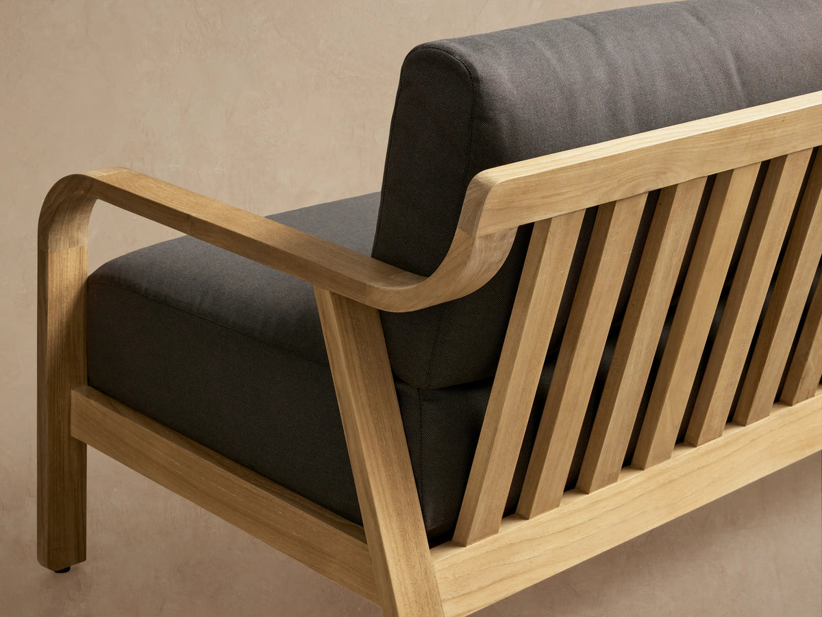 Zoe Outdoor Sofa with light teak wood frame and dark gray cushions, on a light patio floor against a beige wall with shadows.