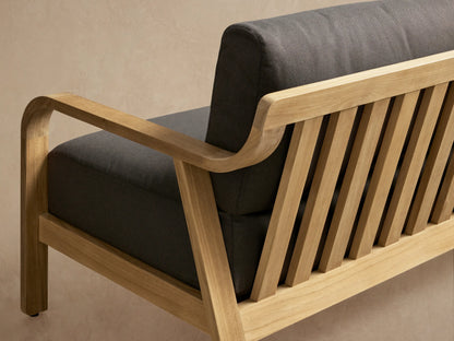 Zoe Outdoor Sofa with light teak wood frame and dark gray cushions, on a light patio floor against a beige wall with shadows.