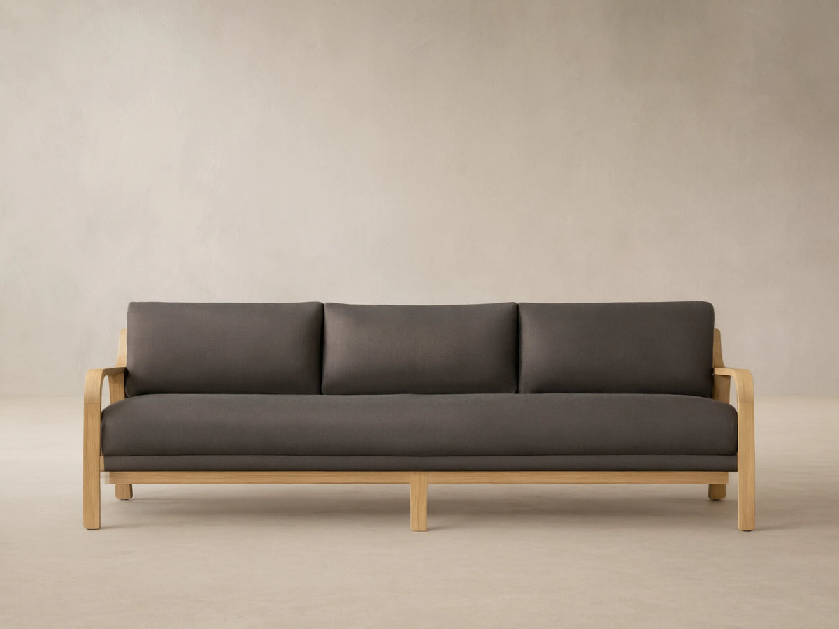 Zoe Outdoor Sofa with a natural teak wood frame, dark gray cushions, 3 back cushions, & 1 seat cushion, on a patio.