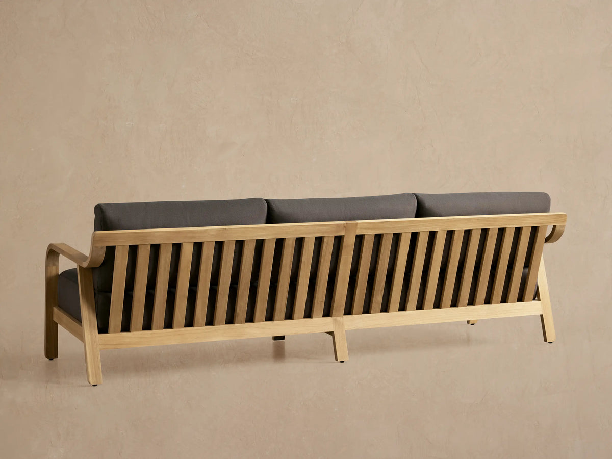 Close-up of Zoe Outdoor Sofa's right side, featuring a slatted natural teak wood frame and dark grey cushions.