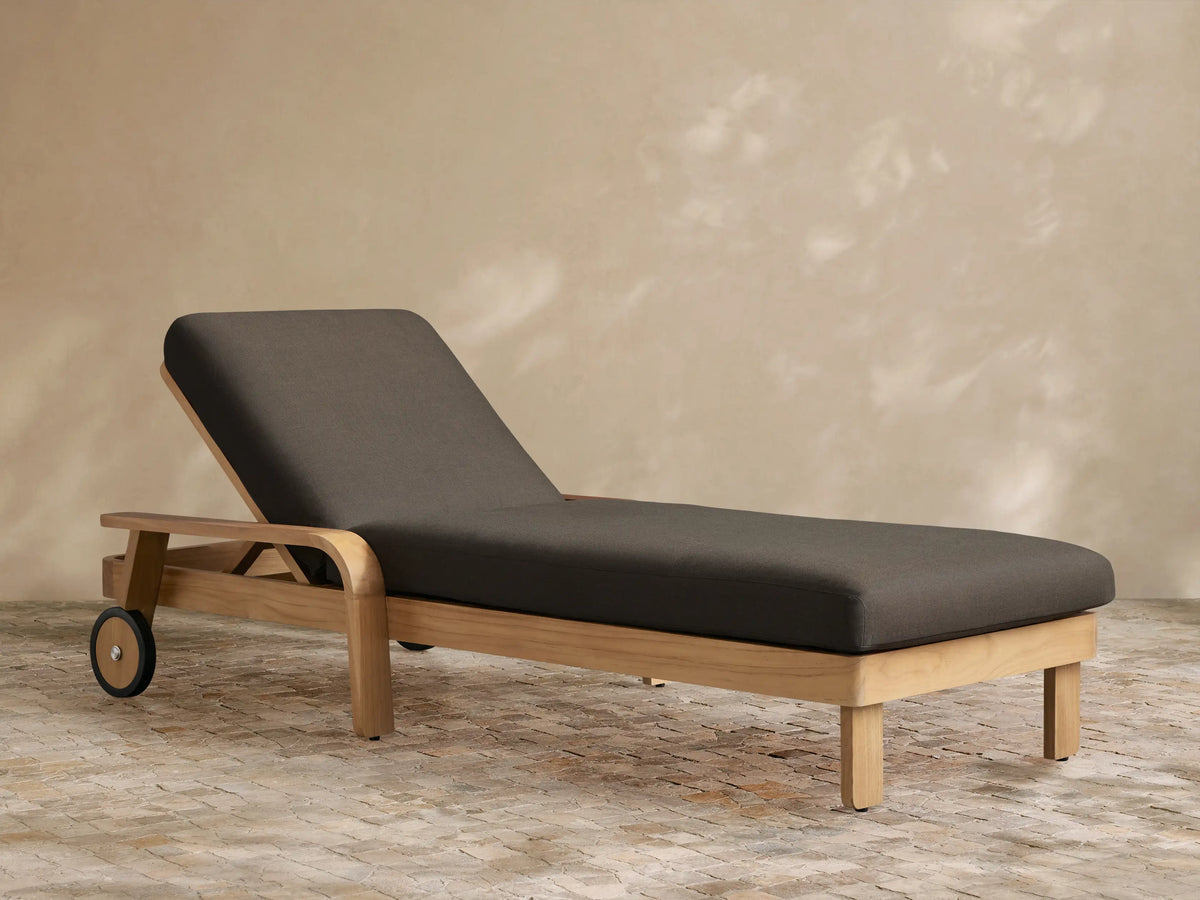 Zoe Outdoor Chaise with teak wood frame, dark gray cushion, and wheels, on a stone patio against a beige wall.