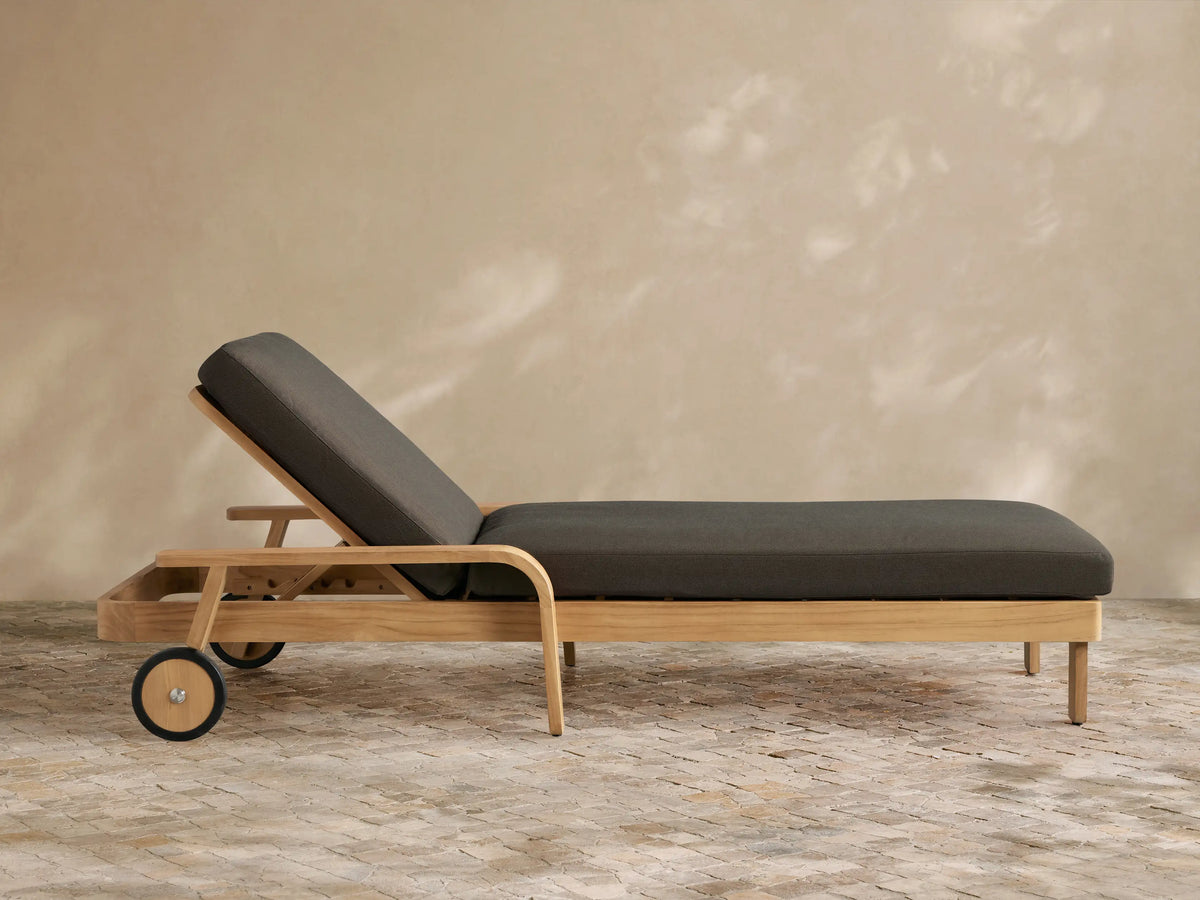 Zoe Outdoor Chaise with teak wood frame, dark gray cushion, and wheels, on a stone patio against a beige wall.