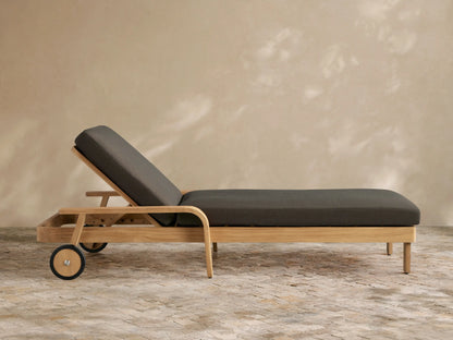 Zoe Outdoor Chaise with teak wood frame, dark gray cushion, and wheels, on a stone patio against a beige wall.