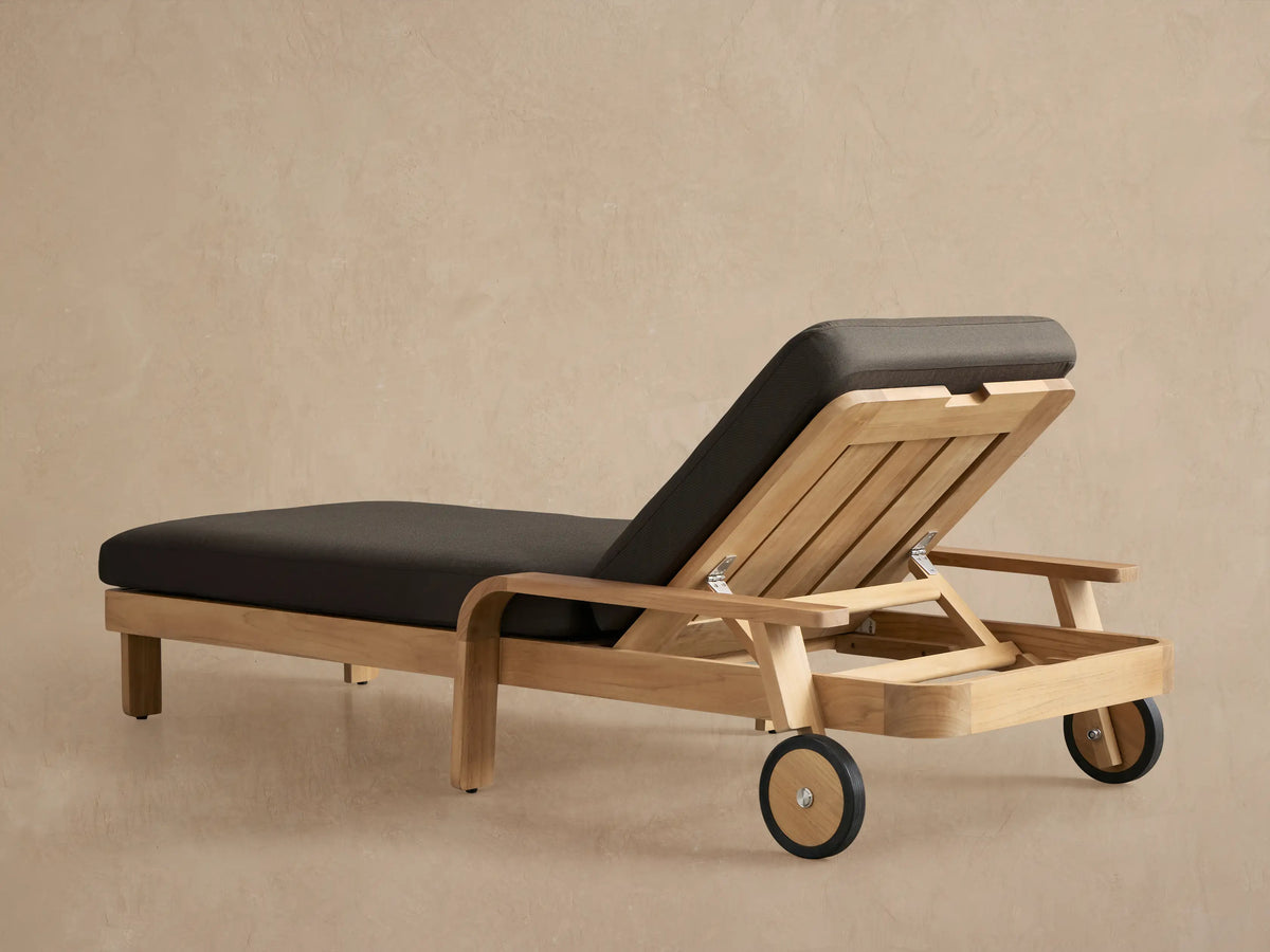 Close-up of a Zoe Outdoor Chaise, light natural teak wood frame with dark gray cushion, showing adjustable back and metal hinge.