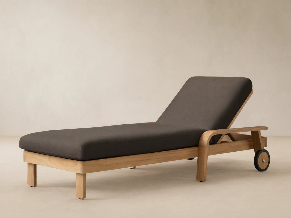 Zoe Outdoor Chaise with teak wood frame, dark gray cushion, and wheels, on a stone patio against a beige wall.